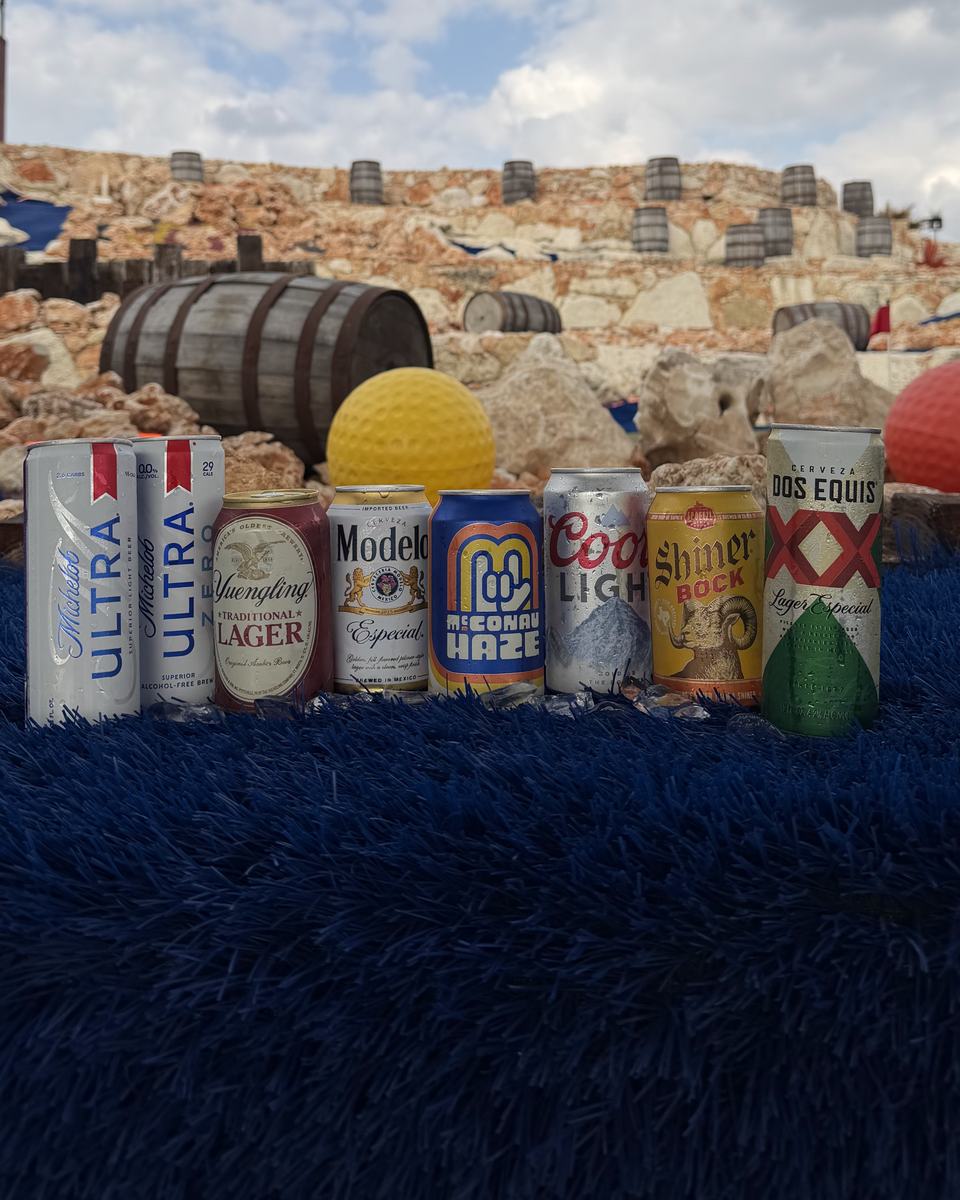 Cans of Michelob Ultra, Modelo, Coors Light, Shiner Bock and other beers lined up on blue turf in front of the rockwork